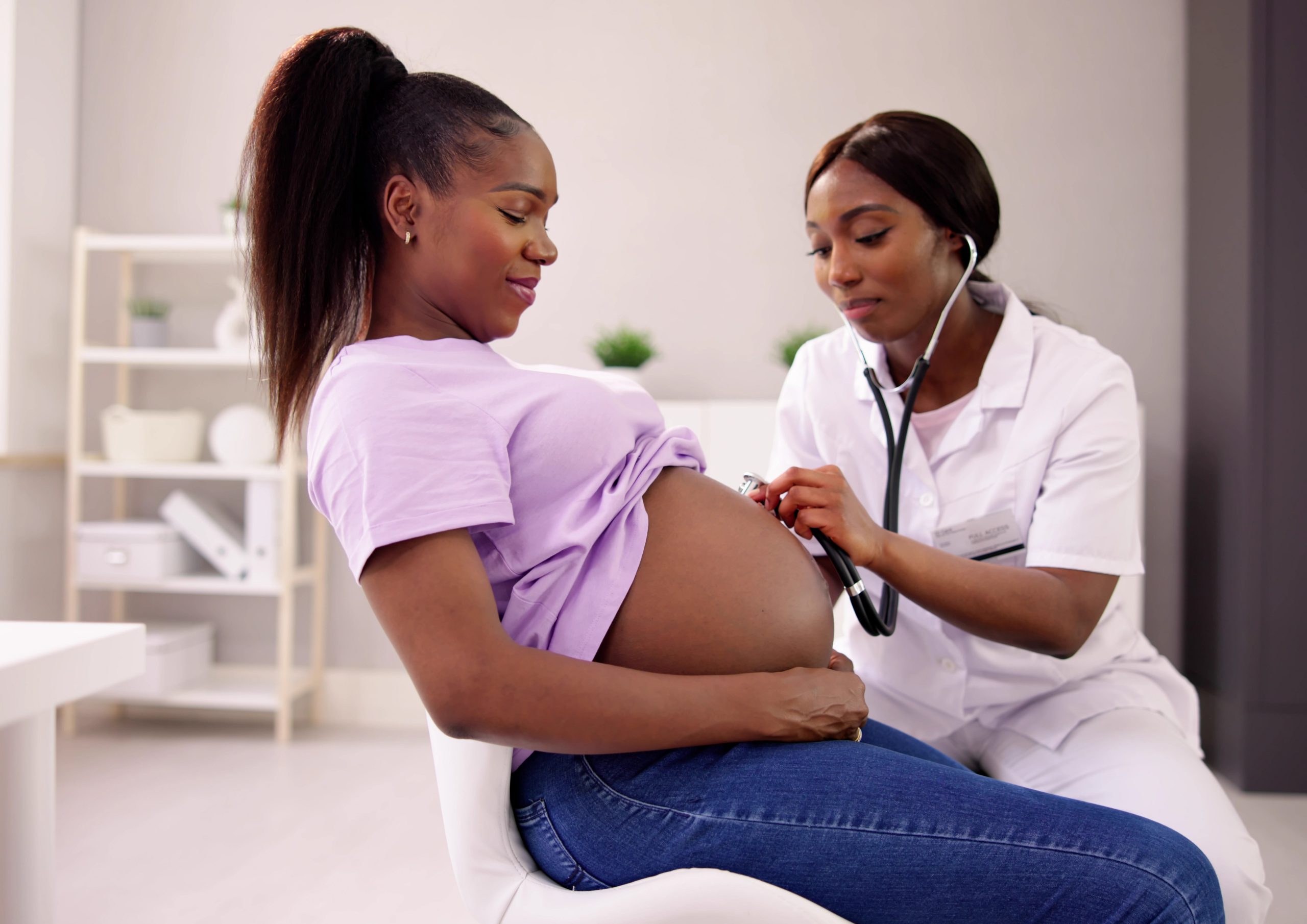 A community health worker uses a stethoscope to check a seated pregnant woman's blood pressure during a home visit.